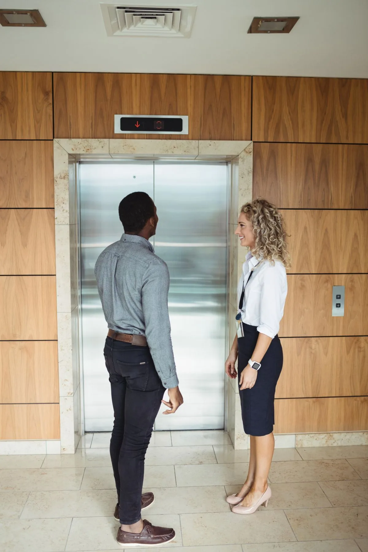 Two people waiting at a stainless steel lift in a timber-panelled lobby